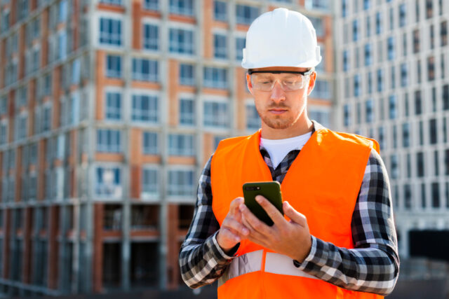 Imagem de um trabalhador da construção civil com EPIs olhando o celular na sua mão