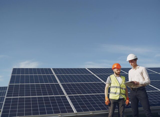 Imagem de um construtor e um engenheiro analisando dados no papel em frente a várias placas de energia solar